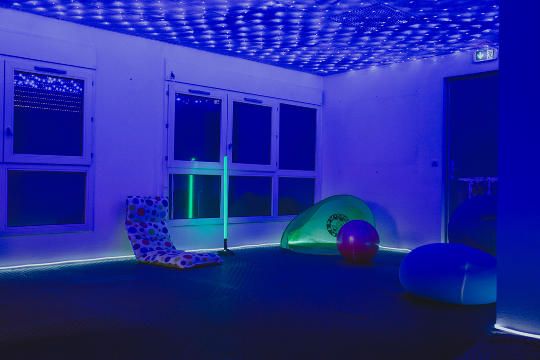 Une salle de l’École inclusive éclairée de lumières bleues, avec un plafond étoilé, des assises au sol, un ballon et une petite tente.